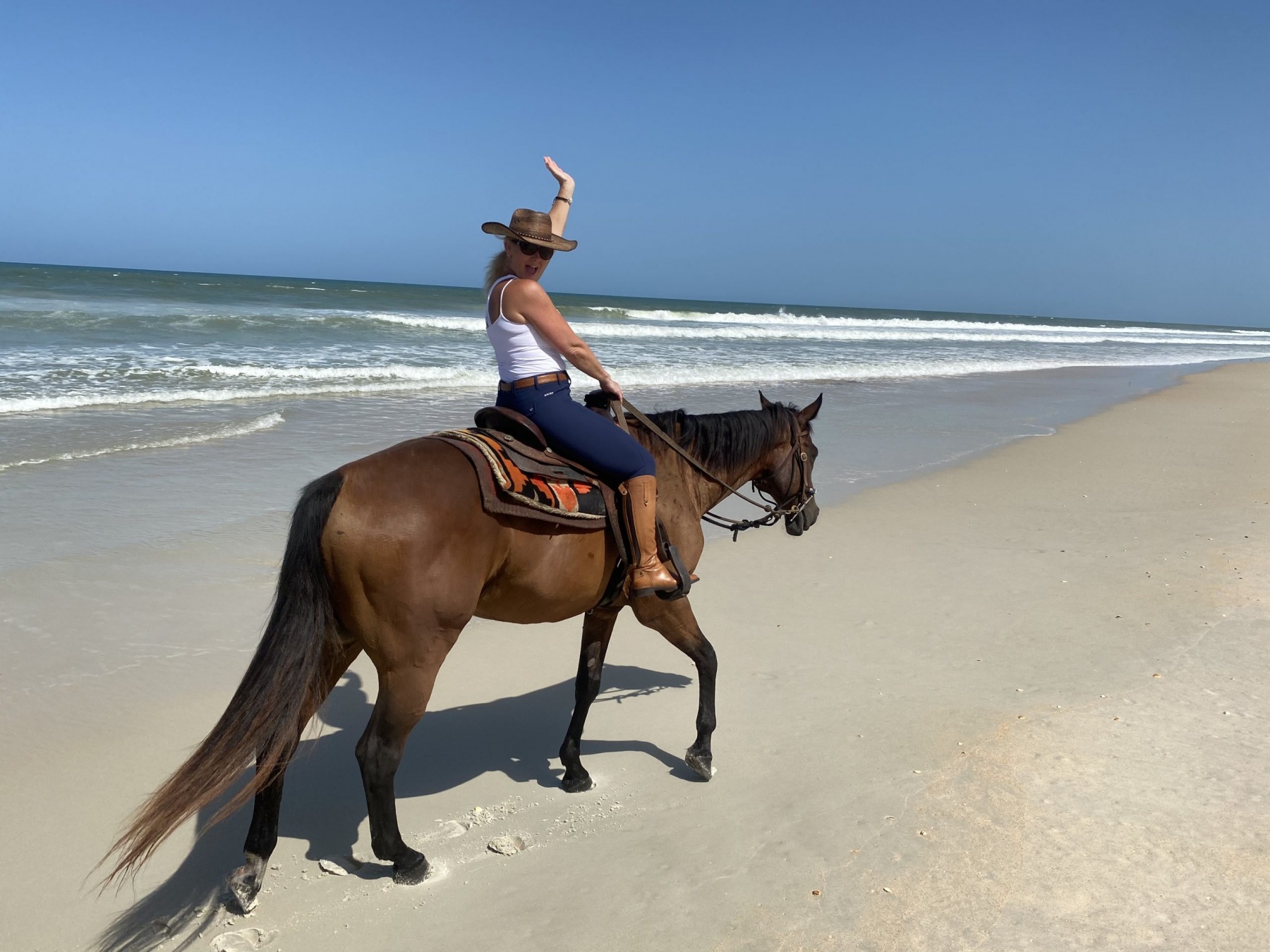A rider waving from a bay horse on the wet sand of the beach
